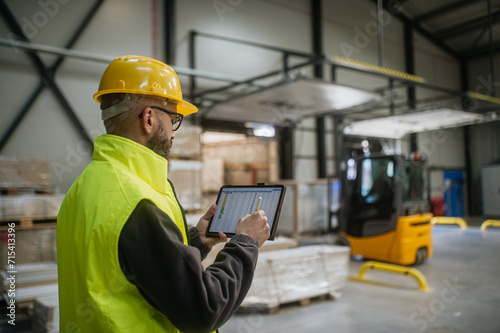 Warehouse receiver standing by delivered cargo, holding tablet, looking at cargo details, checking delivered items, goods against order.