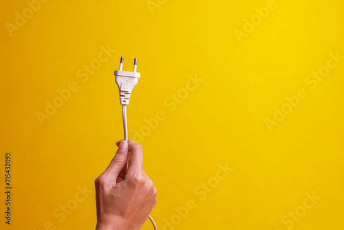 Hand holding white electrical plug isolated on yellow background with copy space. Save energy and earth hour concept