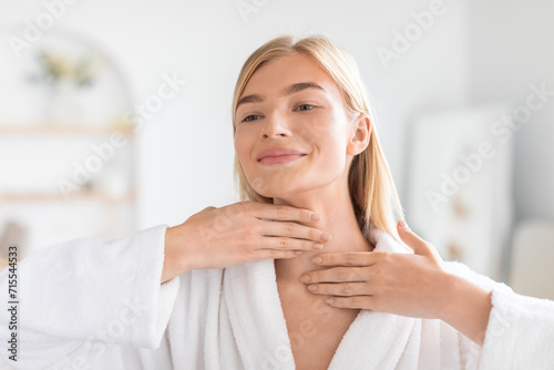 Pretty blonde woman applying moisturizing cream on her neck indoors