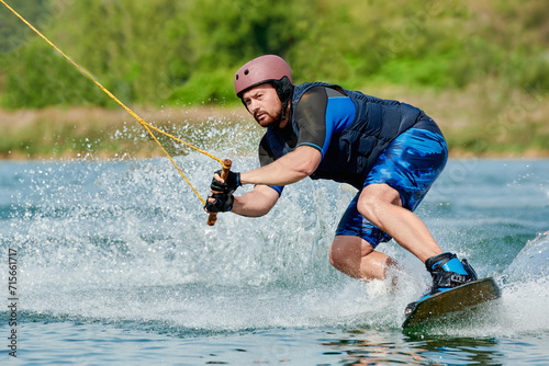 a man on a wakeboard wearing a helmet holds onto a winch and sits down before performing a spray trick