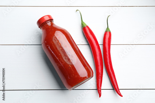 Spicy chili sauce in bottle and peppers on white wooden table, flat lay