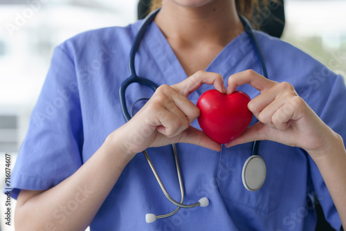A nurse in blue scrubs holds a red heart in her hands, forming a heart shape around it, symbolizing care.