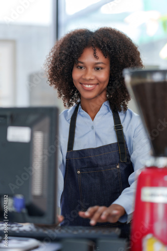 Smiling young barista in apron using cash register at modern cafe.
