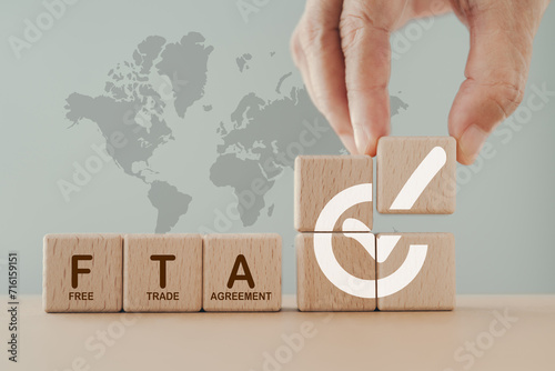 Hand arranged wooden cube blocks with approved signed and word FTA, abbreviation for Free Trade Agreement, and blurred stack of coin on world map background