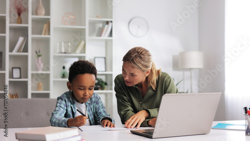 Blonde tutor teaching little African American preschooler to write at home. Help with early childhood education in an online school. Happy boy is learning to write.