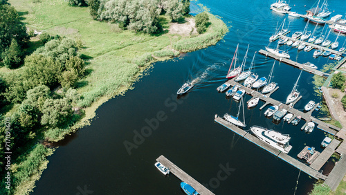 Aerial view of a peaceful marina with various sailboats docked along the pier, surrounded by lush greenery on a bright sunny day