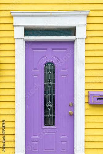 The entrance to a vibrant yellow wooden house with clapboard siding and a purple mailbox. There's a single purple metal door with a narrow glass decorative window, white trim, and a transom window.