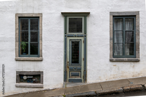 The exterior of a vintage white masonry wall with multiple glass windows, half glass single door, a transom window over the entrance and green colored trim. The facade of the house is on a steep hill.