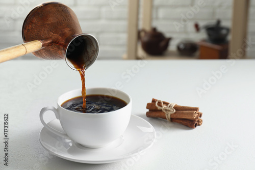 Turkish coffee. Pouring brewed beverage from cezve into cup at white table indoors