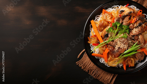 Top view of korean food in black frying pan on dark surface. Cooked asian ingredients in iron skillet on black background with copy space for text. Sautéed noodles, mushrooms and vegetables recipe.