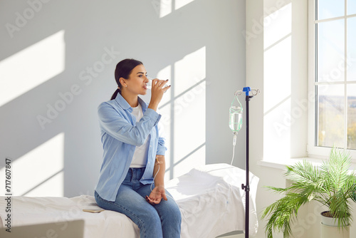 Woman with sterile tube in arm sitting by pole with IV bottle at medical center, drinking water and receiving vitamin therapy infusion to boost body immune system, preserve beauty or fight hangover