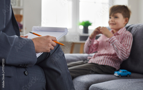 Woman psychologist or therapist with pencil and paper notepad having conversation therapy session with child kid boy who is sitting on sofa with toy car in background. Clipboard, woman's hand close up