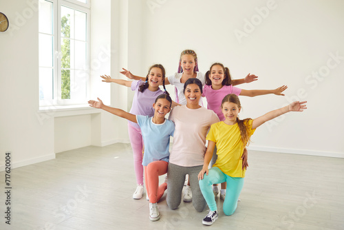 Portrait of cheerful happy active little girls sitting on the floor with their smiling friendly choreographer woman in choreography class, posing and looking at camera after dance workout.