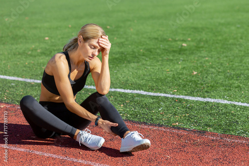 Tired female athlete in sportswear sitting on a running track, holding her head in stress or exhaustion after training.