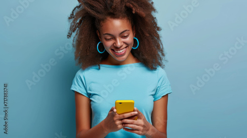smiling young woman looking down at her yellow smartphone with pleasure, wearing a blue top and matching blue hoop earrings against a soft blue background