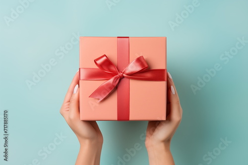 Top view of hands holding craft paper gift box with ribbon bow on isolated pastel blue background. Generated AI.