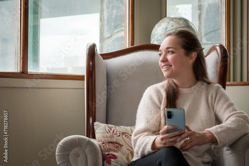 Mujer mirando por la ventana sonriente mientas usa su celular, sentada cómodamente en su sillón