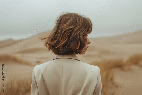 anonymous female with a bob haircut in a cream blazer, back to the camera, against soft dune contours