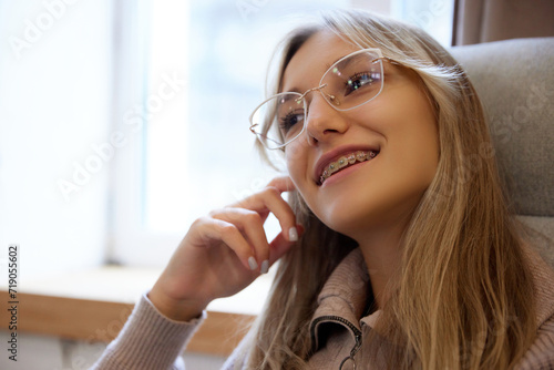 Close up portrait of young woman with dental braces system sitting in armchair near window and dreaming. Concept of beauty and medicine, dental care, malocclusion, orthodontic health. Ad