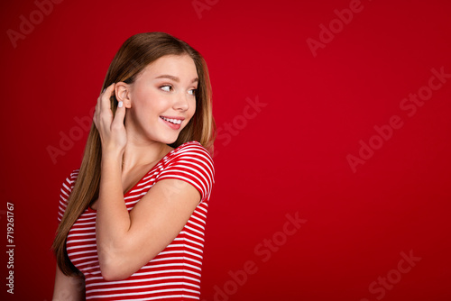 Portrait of adorable teenager wear striped t-shirt tucking hair behind ear look at discount empty space isolated on red color background