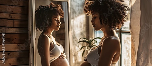 Pretty preggy girl with dark skin and curly hair standing in front of mirror examining her reflection touching her naked belly noticing changes of her body Happy conscious pregnancy. Copy space image