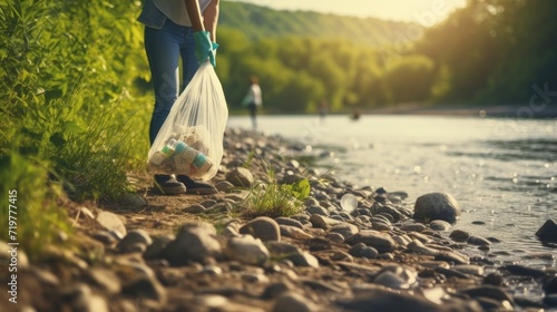 Closeup of a young volunteer holding a trash bag and picking up litter along a riverbank.