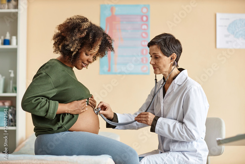 Side view of young pregnant African American woman looking at her big bare belly while senior Caucasian gynecologist checking baby with stethoscope in modern clinic