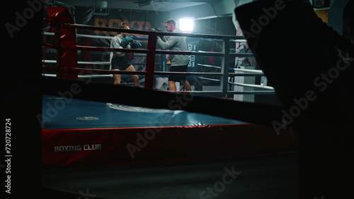 Female boxer in boxing gloves practices fighting technique with male coach and prepares to tournament in dark gym. Athletic fighter hits punching mitts on boxing ring. Physical activity and workout.