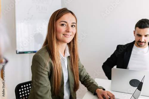 Cheerful young eastern european businesswoman looking at camera sitting with business colleagues in the office. Successful portrait of european professional female worker at workplace.