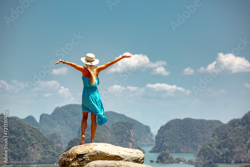 Beautiful young lady in blue dress and white hat is standing with open arms on big rock with sea and islands view. Phang Nga province, Thailand