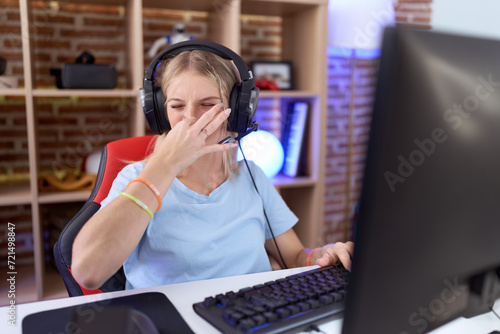 Young caucasian woman playing video games wearing headphones smelling something stinky and disgusting, intolerable smell, holding breath with fingers on nose. bad smell