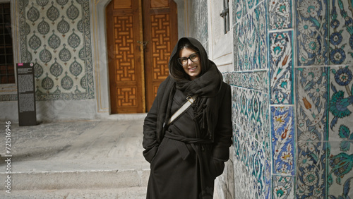 Smiling young woman in a scarf stands by ornate tiles at istanbul's historical topkapi palace.