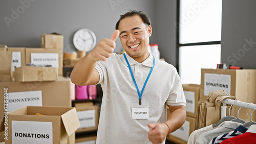 Confident young chinese man, smiling serviceman showcasing the thumb-up gesture in charity center, embodying the spirit of volunteerism and altruism in community services