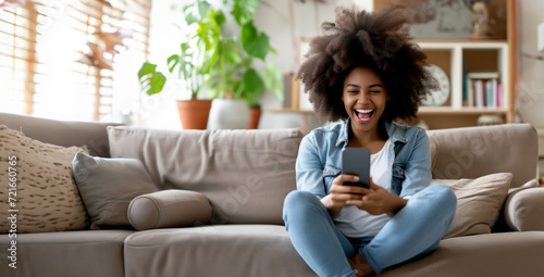 Excited happy young black woman holding smart phone device sitting on sofa at home - Happy satisfied female looking at mobile smartphone screen - Technology concept
