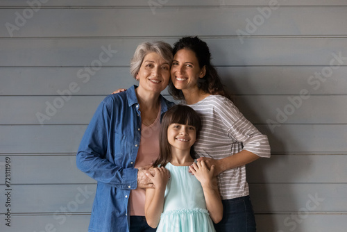 The dearest people. Happy small school age girl daughter her young mom and senior retired grandma look at camera embrace. Three latin female family members stand close look at camera hug for portrait