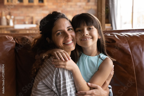 Sweet moment. Headshot portrait single latin mother sit on sofa hug loving little child daughter spend weekend at home together. Caring millennial mom and cute preteen kid girl cuddle look at camera