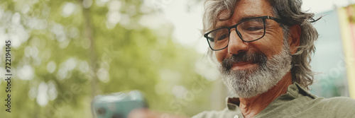 Panorama, Positive middle-aged man with gray hair and beard wearing casual clothes sits on bench and uses mobile phone. Mature gentleman in eyeglasses writes message on smartphone