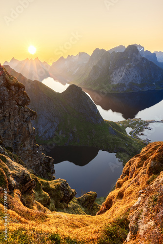 Sunset rays pierce through jagged peaks at Reinebringen, Lofoten Islands, illuminating the mossy foreground and mirrored lakes below, showcasing Norway's midnight sun