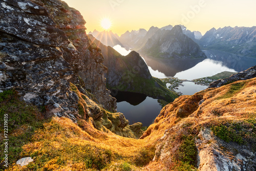 The midnight sun pierces the sky above Reinebringen, illuminating the moss-draped rocks and casting a radiant glow over the dramatic peaks and fjords of Lofoten Island