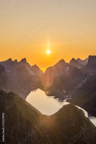 The vertical frame captures the midnight sun casting a warm glow over Reinebringen's sharp peaks in Lofoten, Norway, with the sun's reflection shimmering in the serene fjord