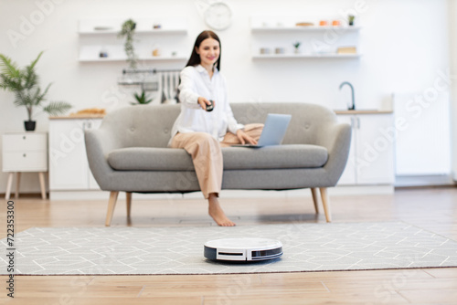 Beautiful lady switching on robot vacuum cleaner via remote control while sitting on sofa in kitchen. Young caucasian brunette woman with laptop programming automatic tidying of room floor.