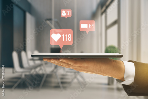 Close up of businessman hand holding smartphone with creative social media icons hologram on blurry office interior background. Community, likes, shares and communication concept.