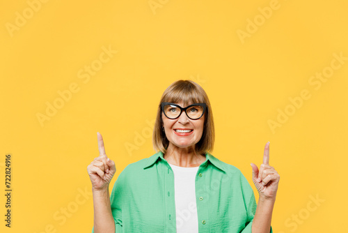Elderly blonde woman 50s year old wear green shirt glasses casual clothes point index finger overhead on area indicate on area copy space mock up isolated on plain yellow background Lifestyle concept