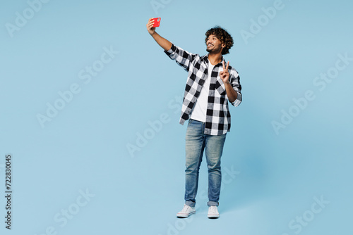 Full body young Indian man wear shirt white t-shirt casual clothes doing selfie shot on mobile cell phone post photo on social network show v-sign isolated on plain blue background. Lifestyle concept