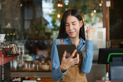 Young businesswoman, owner of an Asian cafe, in navy blue shirt wearing brown apron, is looking tablet. which she held happy smile and delighted by bakery sales of the family's small business cafe.