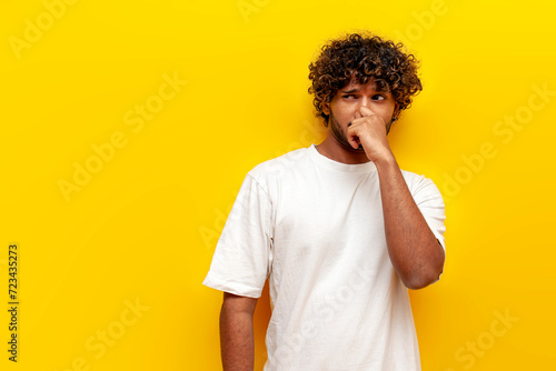 young curly indian man covering his nose with his hand and showing disgust on a yellow isolated background, displeased indian man sniffing an unpleasant smell