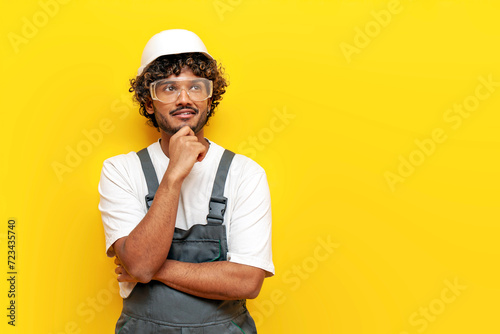 young pensive indian male builder in hard hat and overalls plans and thinks on yellow isolated background, indian foreman in uniform dreams and imagines and looks at copy space