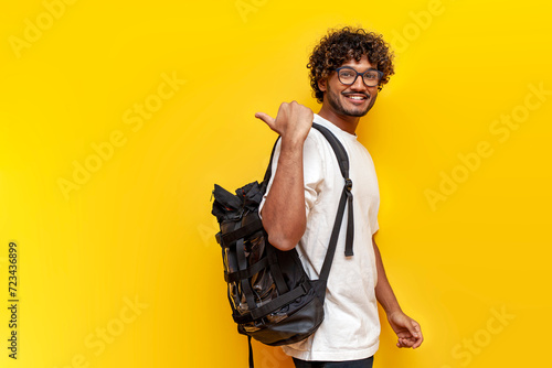 young indian guy student in a white t-shirt with glasses and with backpack points back on yellow isolated background, curly man goes to college to study and points at the copy space from behind