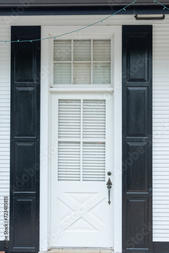 A navy blue wooden house wall with a half glass four-pane window in the panel exterior door. There's a metal doorhandle, transom window with white curtains. The doorway entrance is to a country house.