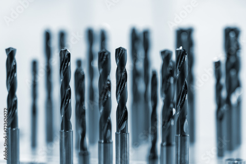 Collection of drill bits in blue hue. They are arranged in two rows in front of white background. Drills are made of metal and have spiral design